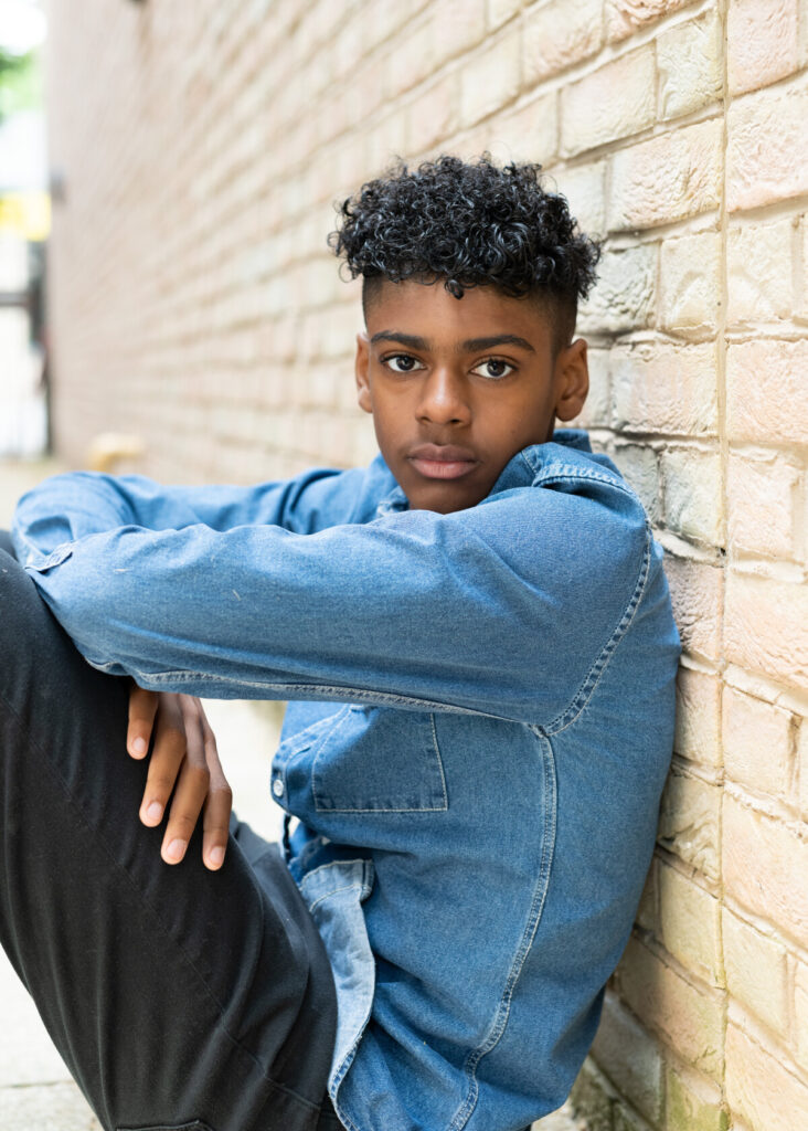A teenage boy sits down against a brick wall with his arms crossed. He has black curly hair and brown eyes and stares into the camera.